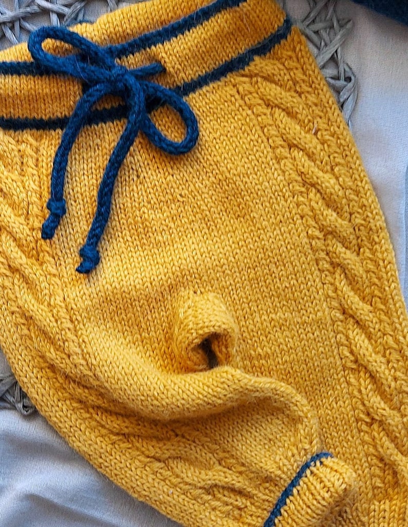 K&ouml;nnte beinhalten: Gelbe gestrickte Babyhose mit blauem Kordelzug und blauem Besatz. Die Hose hat ein Zopfmuster.