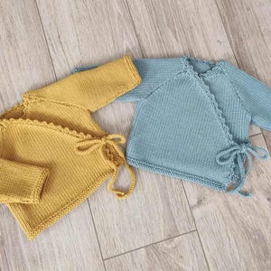 Op de afbeelding: Twee gebreide babywikkelvesten. Eén is mosterdgeel, de andere lichtblauw. Beide hebben lange mouwen en striksluitingen. De vesten zijn gemaakt van zacht garen en geschikt voor pasgeborenen.