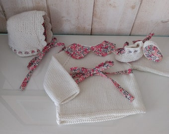 May include: A white knitted baby outfit set featuring a bonnet, sweater, booties, and blanket. The set is accented with floral patterned fabric on the collar, bows, and trim. The outfit is displayed on a white surface.