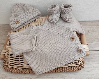 May include: A knitted baby set in a neutral beige color. The set includes a beanie hat, a wrap-style cardigan with wooden buttons, and a pair of booties. The items are displayed on a woven basket, showcasing the handmade quality of the baby clothing.