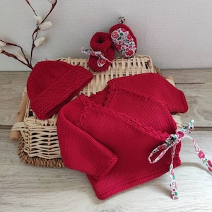 Op de afbeelding: Een rode gebreide babyset met een trui met lange mouwen en een wikkelgesp, een bijpassende muts en een paar slofjes. De trui is vastgebonden met een bloembies.