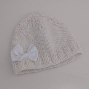 Könnte beinhalten: Eine weiße Strickmütze mit einer weißen Schleife aus Satin. Die Mütze ist mit einem einfachen Stichmuster gearbeitet und hat einen umgeschlagenen Rand.