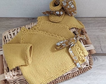 May include: A mustard yellow knitted baby sweater with long sleeves and a wrap style closure. The sweater is tied with a floral ribbon and is displayed in a wicker basket.