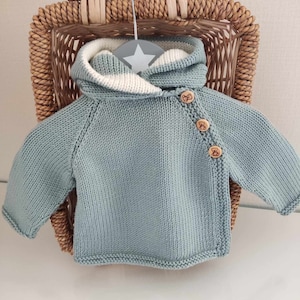 Op de afbeelding: Een lichtblauwe gebreide babytrui met een witte capuchon en houten knopen. De trui hangt aan een hanger in een rieten mand.