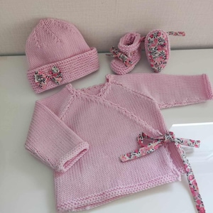 Op de afbeelding: Een roze gebreide baby-outfitset met een muts, trui en slofjes. De trui heeft een wikkel-stijl met een bloemenlint. De slofjes hebben bloemenstof accenten.