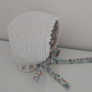 May include: White knitted baby bonnet with a floral print lining and matching ribbon ties.