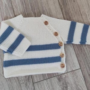 Op de afbeelding: Een gebreid babyvestje met crème en blauwe strepen. Het vestje heeft lange mouwen en houten knopen. De blauwe strepen zijn horizontaal. Het kledingstuk is gemaakt van zacht, getextureerd garen.