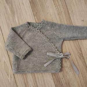 Op de afbeelding: Een lichtgrijze gebreide babywikkelvest met lange mouwen. Het vest heeft een geschulpte rand en een sluiting met een ruitjeslint. Het kledingstuk is gemaakt van zacht, getextureerd garen en ligt op een houten oppervlak.