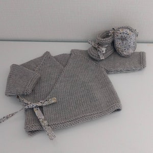 May include: A gray knitted baby sweater with a wrap-style closure and a floral ribbon tie. The sweater is paired with a set of gray knitted baby booties with floral fabric accents.