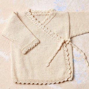 Op de afbeelding: Crèmekleurige gebreide babywikkel trui met een geschulpte rand en striksluiting. De trui heeft een V-hals en lange mouwen. Het breiwerk is gemaakt van zachte, natuurlijke vezels, perfect voor pasgeborenen en baby's.
