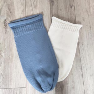 Può includere: Due calzini per neonati lavorati a maglia, uno in un azzurro tenue e l'altro bianco, sono esposti su una superficie di legno chiaro. I calzini hanno un polsino a coste e una punta arrotondata, suggerendo un design confortevole.