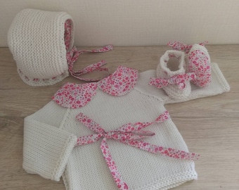 May include: A white knitted baby outfit with a pink floral trim. The outfit includes a hat, sweater, and booties.