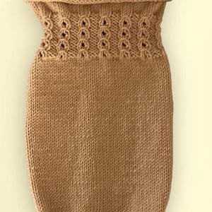 May include: A tan-colored knitted baby cocoon. The cocoon features a textured pattern around the top with small circular openings. The body of the cocoon is a simple knit stitch. The bottom of the cocoon is gathered with a small tie.