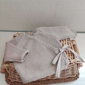 Op de afbeelding: Lichtbeige gebreide baby set bestaande uit een trui en bijpassende muts. De trui heeft lange mouwen en een wikkelontwerp, vastgezet met een geruite strik. De set wordt gepresenteerd op een rieten mand.