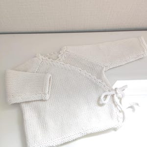 Op de afbeelding: Een witte, gebreide babywikkelvest met lange mouwen. Het wikkelontwerp heeft striksluitingen aan de zijkant. De halslijn en randen hebben een delicate, geschulpte afwerking. Het kledingstuk is gemaakt van zacht, getextureerd garen.