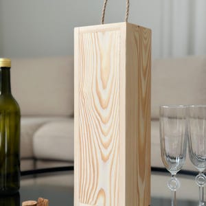 May include: A natural wood wine bottle gift box with a rope handle. The box is made of light wood with a natural finish.