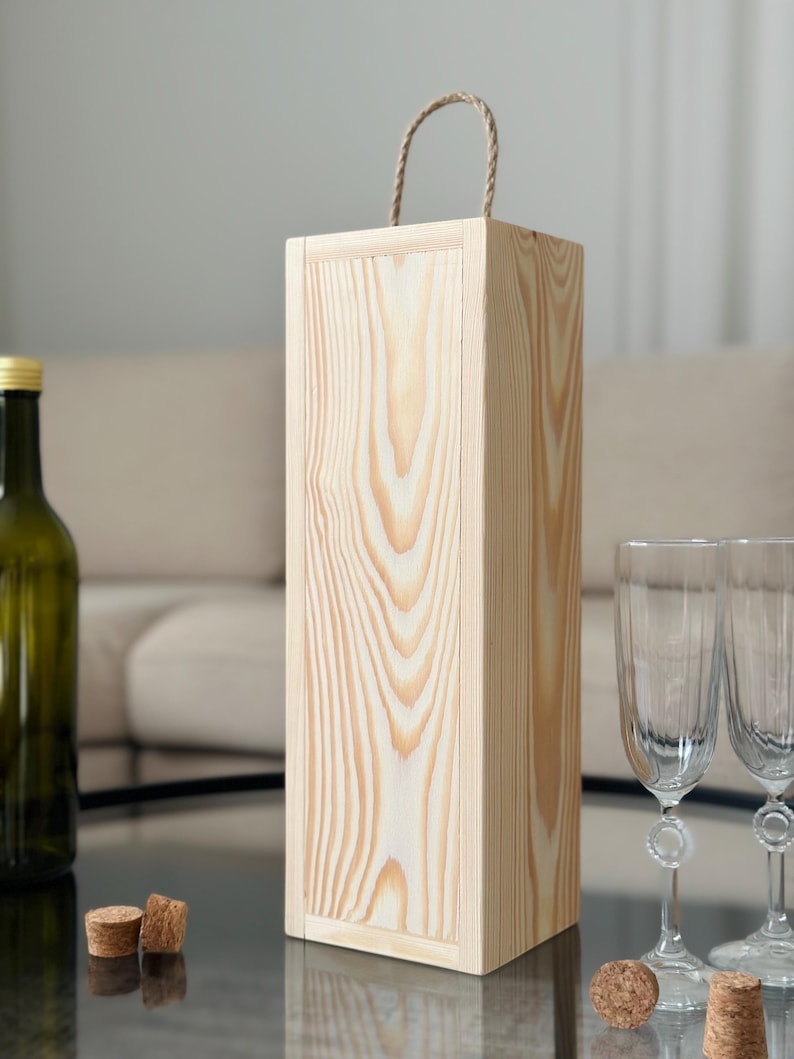 May include: A natural wood wine bottle gift box with a rope handle. The box is made of light wood with a natural finish.