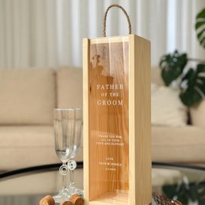 May include: Wooden box with a clear glass front, designed to hold a bottle of wine. The box has a rope handle and is engraved with the text "Father of the Groom", "Thank you for all of your love and support", "Love Zach & Jessica 3.1.2025".