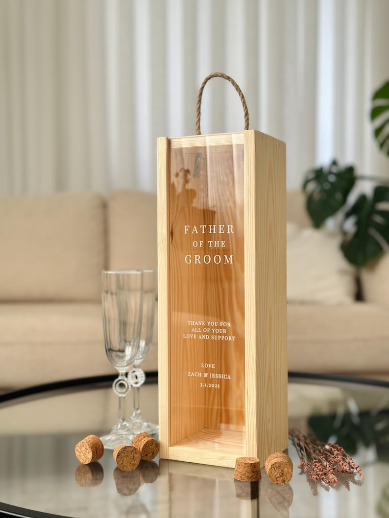 May include: Wooden box with a clear glass front, designed to hold a bottle of wine. The box has a rope handle and is engraved with the text "Father of the Groom", "Thank you for all of your love and support", "Love Zach & Jessica 3.1.2025".