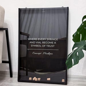 May include: A black framed sign with the text "WHERE EVERY SYRINGE AND VIAL BECOME A SYMBOL OF TRUST" and "Concept Medspa" in white. Several corks are at the bottom of the frame. A monstera plant is in the background.