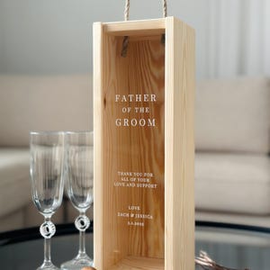 May include: Wooden box with a rope handle, designed for a bottle of wine. The box has a clear window and is engraved with the text "Father of the Groom", "Thank you for all of your love and support", "Love Zach & Jessica 3.1.2025".
