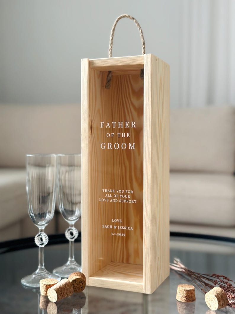 May include: Wooden box with a rope handle, designed for a bottle of wine. The box has a clear window and is engraved with the text "Father of the Groom", "Thank you for all of your love and support", "Love Zach & Jessica 3.1.2025".
