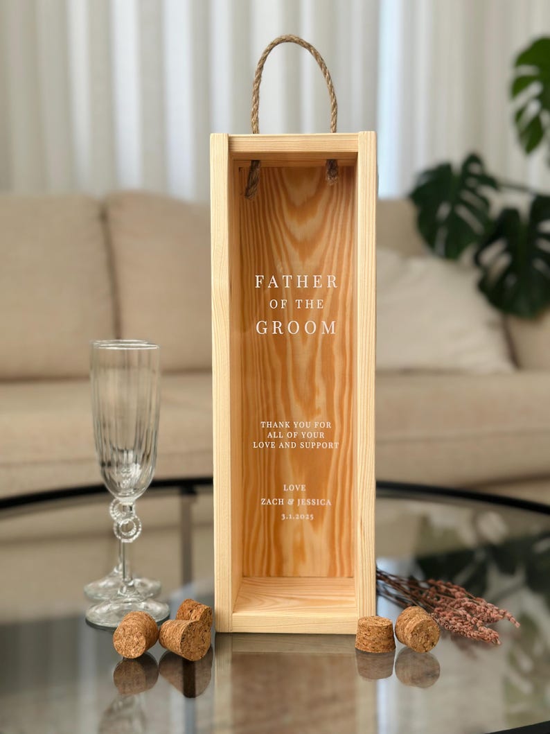 May include: Wooden box with a rope handle, designed for a bottle of wine. The box is engraved with the text "Father of the Groom", a message of thanks, and the names "Zach & Jessica" with a date.