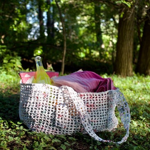 May include: A white crocheted tote bag with a pink and white striped towel and a clear glass bottle inside. The bag is sitting on a bed of green grass.