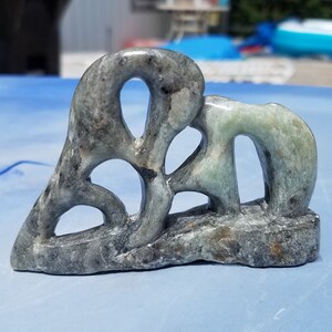 May include: A carved stone sculpture with a gray and green color scheme. The abstract design features open spaces and rounded shapes, resting on a rectangular base. The sculpture is displayed on a blue surface.