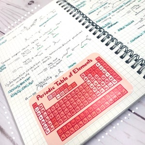 May include: A pink and red periodic table of elements sticker with the words "Periodic Table of Elements" at the top. The sticker is on a white gridded notebook.