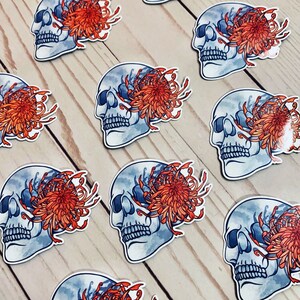 May include: A set of nine stickers featuring a skull with a chrysanthemum flower covering one eye. The skulls are blue and white with a watercolor effect. The flowers are orange and red.