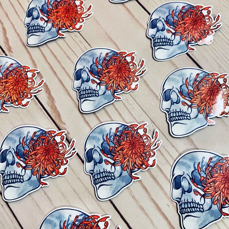 May include: A set of nine stickers featuring a skull with a chrysanthemum flower covering one eye. The skulls are blue and white with a watercolor effect. The flowers are orange and red.