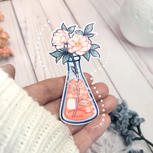 May include: A white flower bouquet in a clear glass beaker with a pink liquid. The beaker has markings for 250 ml, 150 ml, and 100 ml. The flowers are white with pink centers.