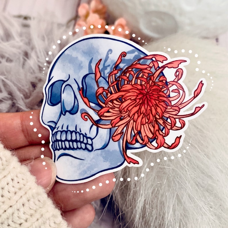 May include: A blue and white skull sticker with a red and pink chrysanthemum flower. The sticker is on a white fuzzy surface.