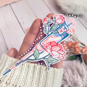 May include: A sticker featuring a pink and blue syringe with a pink flower and green leaves. The syringe is surrounded by a white dotted circle.