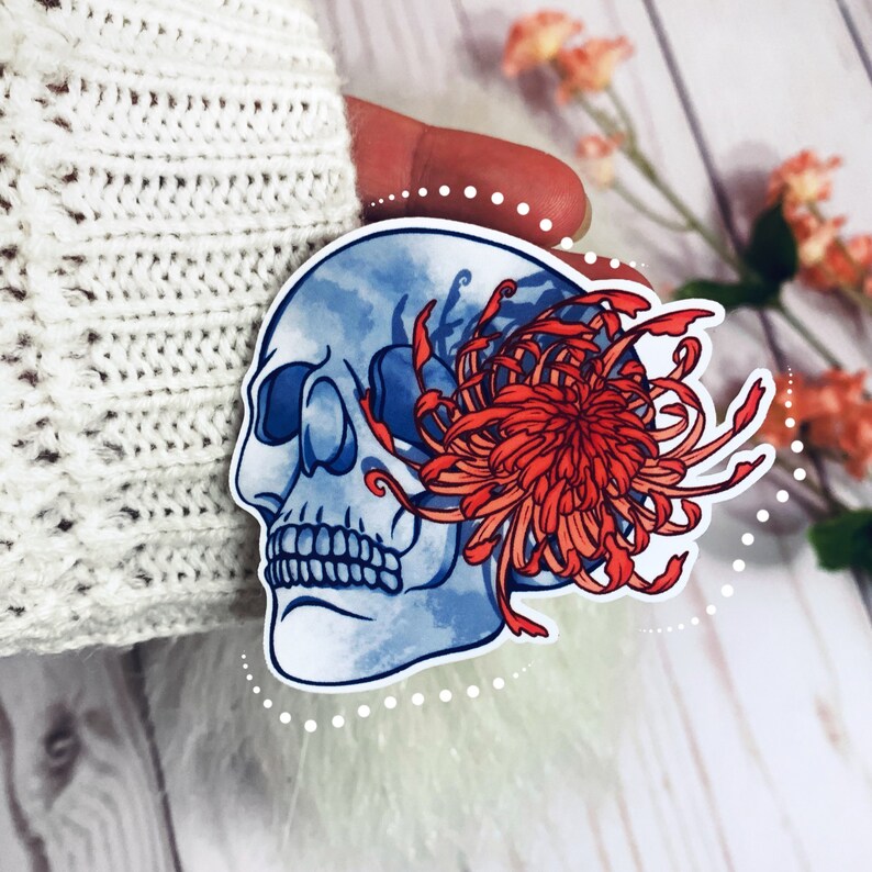 May include: A blue and white skull with a red and orange chrysanthemum flower growing out of its eye socket. The skull and flower are on a white sticker.