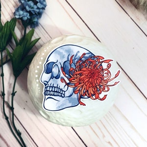 May include: A sticker featuring a blue and white skull with a red and orange chrysanthemum flower. The sticker is on a white background.