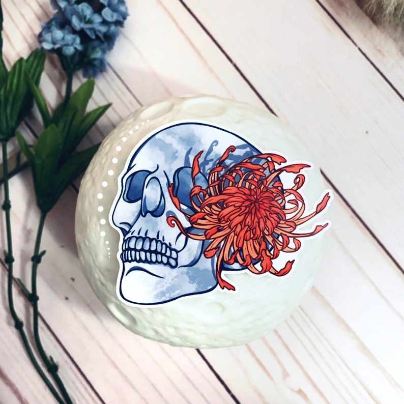 May include: A sticker featuring a blue and white skull with a red and orange chrysanthemum flower. The sticker is on a white background.