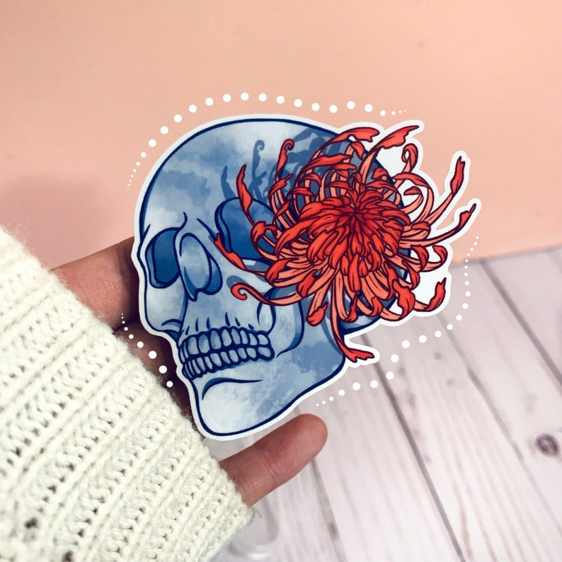 May include: A blue and white skull sticker with a red and orange chrysanthemum flower. The sticker is on a white wooden surface.