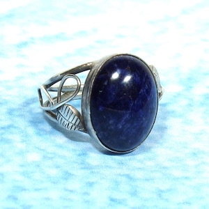 May include: A silver ring featuring a large, oval-shaped, deep blue stone set in a smooth bezel. The band is crafted with openwork metal detailing that includes small, etched leaf-like shapes on either side of the central stone setting.