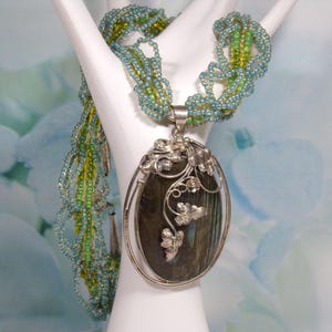 May include: A silver necklace with a large oval pendant featuring a dark green gemstone surrounded by silver floral details. The necklace is made of multiple strands of small blue and green beads.