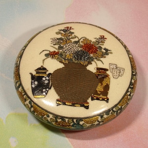 May include: A round, cream-colored ceramic lidded box featuring a central illustration of a brown textured vase filled with colorful chrysanthemums, flanked by a black teapot and a brown vase, with small cups and a floral border.