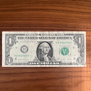 May include: A one dollar bill featuring a portrait of George Washington. The bill is green and has the text "THE UNITED STATES OF AMERICA" and "ONE DOLLAR" printed on it. The serial number is H 03456654.