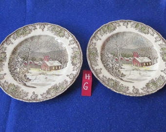 May include: Two white ceramic dinner plates with a winter scene featuring a red schoolhouse and snow-covered trees. The plates have a brown and green leafy border.