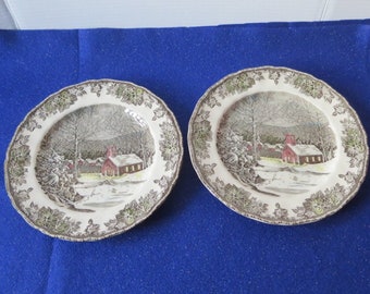 May include: Two vintage ceramic dinner plates with a winter scene of a red schoolhouse in a snowy landscape. The plates have a brown and green leafy border.
