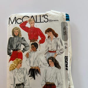 以下が含まれることがあります： McCall'sのブラウス用型紙封筒、番号8297、サイズ14。表面には、長袖、プリーツ、襟やリボンを含む様々なネックラインを備えた5種類のブラウススタイルのイラストが描かれており、スカートやベルトと組み合わされています。