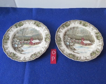 May include: Two white ceramic dinner plates with a winter scene featuring a red schoolhouse and snow-covered trees. The plates have a green and brown leafy border.