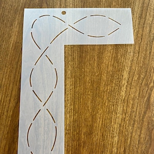 May include: White plastic quilting template with a design of a continuous line of overlapping ovals. The template is shaped like a right angle.