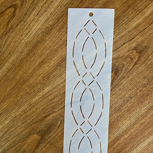 May include: A rectangular stencil with a repeating geometric pattern of connected ovals and diamonds. The stencil is made of a light-colored, semi-transparent material and has a small hole at the top for hanging. The design is cut out, creating negative space.