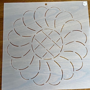 May include: White plastic quilting stencil with a circular design featuring a central cross shape and a ring of curved lines. The stencil is labeled #136.